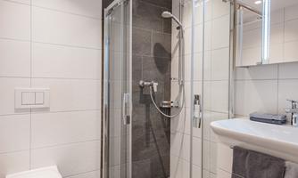 A modern bathroom with a glass shower and a sink. The walls are light-tiled, and there is a toilet in the room.
