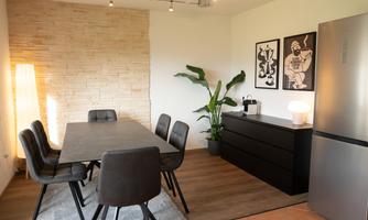 A modern dining room with a large table and six chairs. On the wall hang two artworks, and a plant adds to the cozy atmosphere.