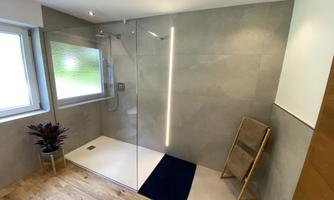 A modern bathroom with a glass shower and tiled walls. A wooden chair and a plant add a cozy touch to the space.