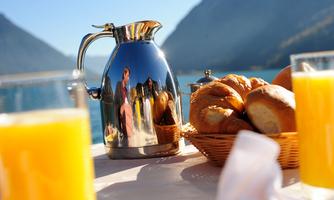 An elegant jug stands on a table by the water, surrounded by fresh bread and glasses of orange juice. In the background, there are mountains and a clear sky.