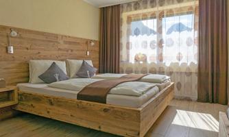 A cozy bedroom with a wooden bed and two pillows. Large windows provide plenty of daylight and a view of the scenery.