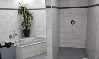 A modern bathroom with white tiles and a spacious shower. A green plant stands in the corner, and the floor is in a light gray tone.