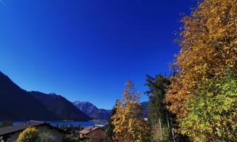 A picturesque view of a lake surrounded by mountains and colorful autumn trees. The clear blue sky gives the landscape a serene atmosphere.