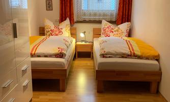 A cozy bedroom with two single beds and colorful pillows. The room has bright walls and wooden flooring, as well as a large window cut-out with a curtain.