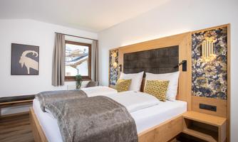 A modern bedroom with a large bed and stylish cushions. The walls are decorated with wood and floral accents.