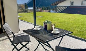 A cozy outdoor area with a table and two chairs. In the background, green meadows and mountains can be seen.