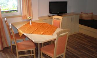 A cozy dining area with a wooden table and orange chairs. A TV is mounted on the wall, and underneath it is a dark seating area.