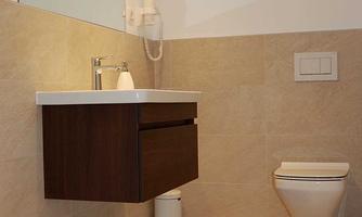 A modern bathroom with a wall-mounted sink and a toilet. The floor is brightly tiled and a brown rug is laid in front of the sink.