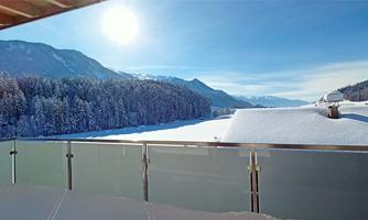 A snow-covered landscape with mountains in the background and a clear blue sky. The terrace offers a beautiful view of the snowy river.