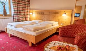 A cozy bedroom with a wooden double bed and a red carpet. In the corner, there is an armchair and a bowl of apples on a side table.