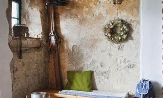 A rustic room with a wooden bench and a green cushion. A wreath hangs on the wall, and a milk can is on the floor.