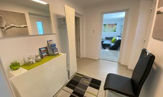 A modern hallway with a bright and minimalist design. On the left side, there is a white cabinet with informational brochures and a black chair on a patterned carpet.