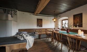 A cozy room with wooden furniture and a large dining table. Against one wall, there is a single bed, and warm lighting is provided by a pendant lamp.