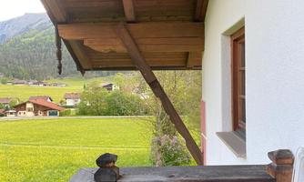 A balcony with a wooden railing and a view of green meadows. In the background, mountains and a few houses are visible.
