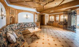 A cozy room with a large, floral-patterned bench and a wooden table. The walls are designed with wooden paneling and decorative lights.
