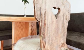 A rustic wooden chair with a heart-shaped cutout and a soft fur seat cushion. In the background, there is a wooden table with a vase and a flower.