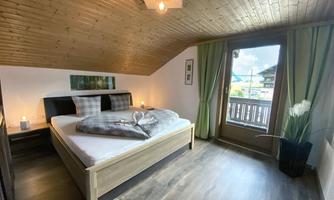 A cozy bedroom with a double bed and wooden ceiling. Large windows let in plenty of light and provide a view outside.