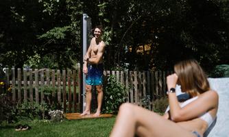 A man is showering in the garden while a woman is relaxing in the sun. The surroundings are green and inviting.