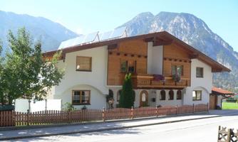 A beautiful, modern house with wooden elements and a balcony. In the background, high mountains and a blue sky can be seen.