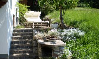 An idyllic outdoor area with a sun lounger and blooming plants. Next to the stairs is a small stone grill area.
