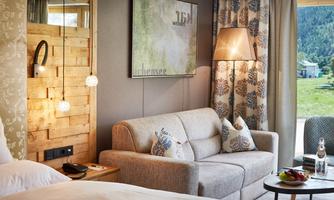 A cozy hotel room with a beige sofa, warm lighting, and a view of nature. The walls are tastefully decorated and there is fresh fruit on the table.