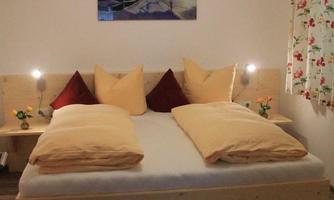 A cozy bedroom with a large double bed and many pillows. A picture hangs on the wall and the windows are decorated with blooming curtains.
