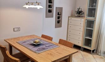 A cozy dining area with a wooden table and four chairs. On the wall, there is a shelf with decorative objects and a large window front.