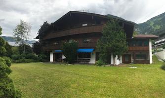 A modern wooden building with a large garden. The sky is cloudy, and the surroundings are green and mountainous.