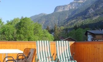 A terrace with two loungers and a table, surrounded by a beautiful mountain panorama. The landscape is green and sunny, ideal for relaxing.