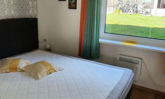 A simple bedroom with a double bed and colorful curtains. A green meadow is visible through the window.