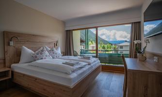 A cozy bedroom with a double bed and light wooden furniture. The view from the window shows a picturesque mountain landscape.