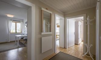 A bright hallway with wooden flooring and white wall paint. There is a mirror as well as doors leading to another room and a coat rack.