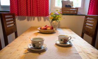 A cozy table with two cups, a fruit bowl, and a vase of flowers. Red curtains and wooden chairs create an inviting atmosphere.