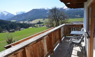 A balcony with a wooden railing offers a beautiful view of green meadows and snow-capped mountains. The sunny landscape conveys a calm and relaxing atmosphere.