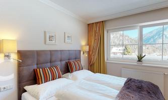 A cozy bedroom with a double bed and colorful pillows. Large windows provide a view of the mountains and let in plenty of light.
