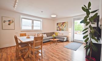 A bright and modern living room with a sofa, a table, and beautiful plants. Large windows provide plenty of light and a pleasant atmosphere.