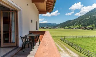 A spacious balcony with a view of a green meadow and mountains behind a modern building. The sky is blue with some white clouds.