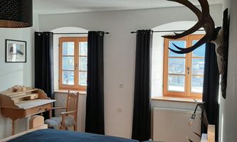 A cozy bedroom with two beds and an antique desk. Large windows provide plenty of daylight and a rustic atmosphere.