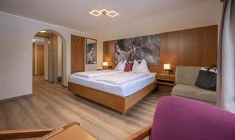 A modern hotel room with a double bed, a cozy armchair, and warm wooden elements. There is a pleasant atmosphere with soft lighting and wall decoration.
