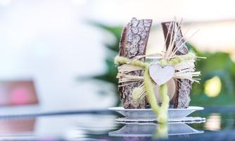 A decorative wooden sculpture with jute ribbon and heart pendant. The background is blurred, featuring green plants.