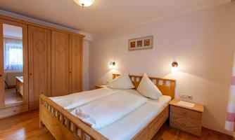 A cozy bedroom with a wooden bed and two pillows. The room has a warm wooden floor and a mirror in a wardrobe.