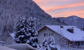 A snowy landscape with snow-covered trees and a house in the foreground. The sky glows in soft shades of pink and purple at sunset.