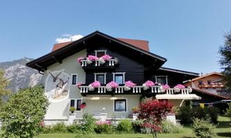 A cozy building with colorful flower balconies amidst a green landscape. In the background, mountains are visible and the sky is clear and blue.