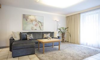 A cozy living room with a gray sofa and a wooden table. Large windows let in plenty of light and a plant adds a touch of nature.