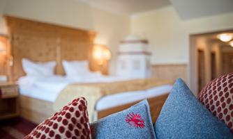 A cozy hotel room with a large bed and warm wooden elements. Decorative cushions are visible in the foreground.