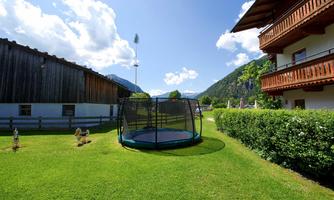 A garden with a trampoline surrounded by green grass. In the background, there are mountains and a bright blue sky.