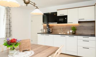A modern kitchen with white cabinets and wooden details. A dining area with a table and a blooming plant is also visible.