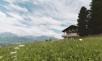 A charming house on a hill, surrounded by meadows and mountains. The sky is overcast, giving the scene a tranquil atmosphere.