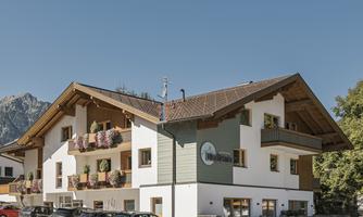 A modern building in a mountainous setting with traditional wooden elements. The facade is bright and welcoming, surrounded by parking lots and nature.