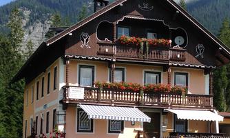 A charming, traditional country house with balconies and blooming plants. In the background, mountains and a clear sky can be seen.